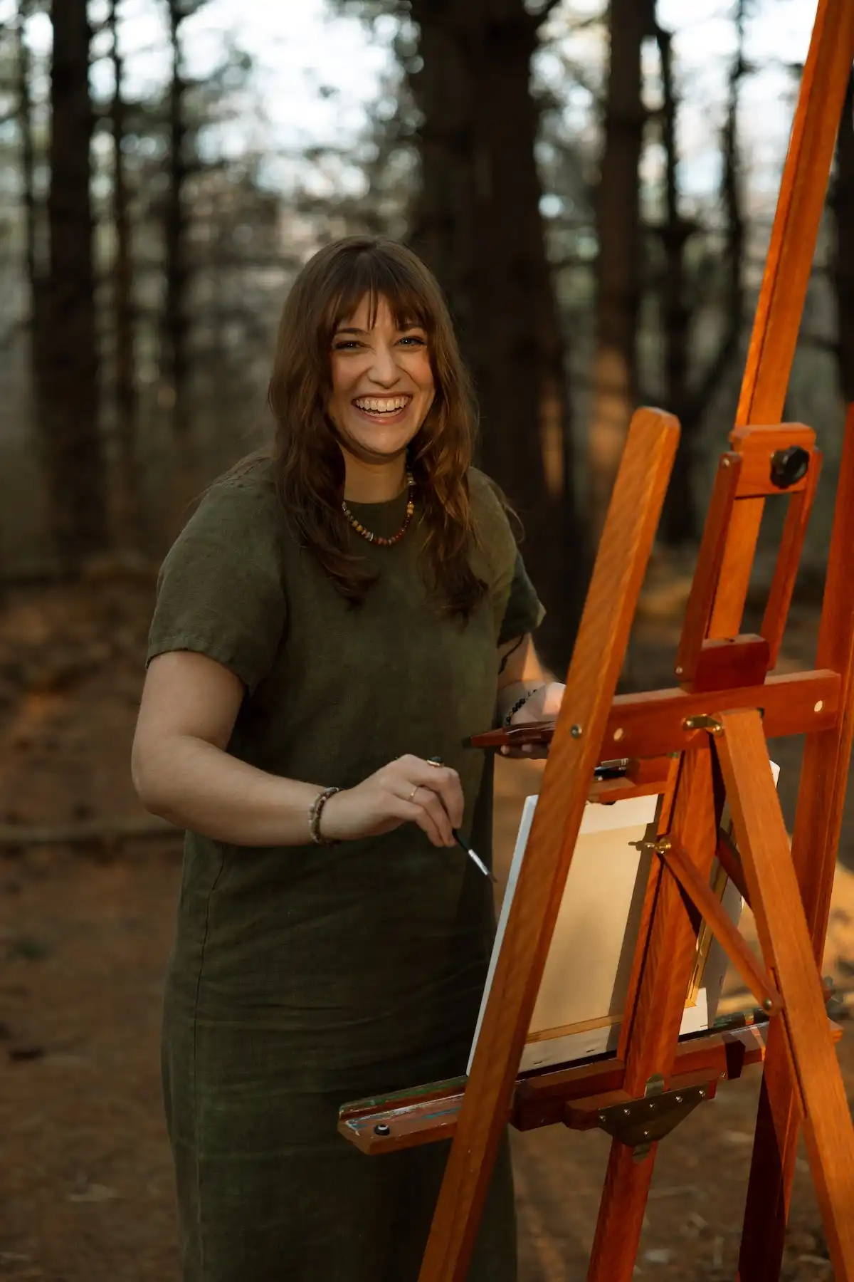 Portrait of Madeline Holloway, credentialed art therapist, painting at an easel in a pine forest